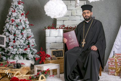 الأب نكتاريوس فرح جالسًا بثوبه الكهنوتي ويحمل كتاب الإنجيل